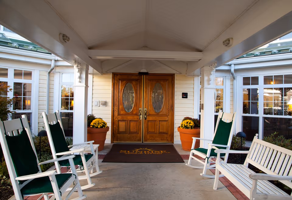 Covered entrance to a senior living facility with double wooden doors featuring oval glass panels. There are white rocking chairs with green cushions and a white bench on either side of the entrance. Large potted plants with yellow flowers flank the doors, and windows with white frames are visible on both sides.
