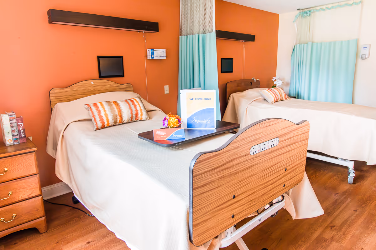 A bright and clean room with two hospital-style beds, each with a wooden headboard and footboard. Both beds have beige bedspreads and decorative pillows with orange and white patterns. A bedside table with drawers is next to one bed, holding some books. A tray with a welcome book and flowers is placed on one of the beds. The walls are painted orange and white, and there are teal privacy curtains between the beds and on the window.