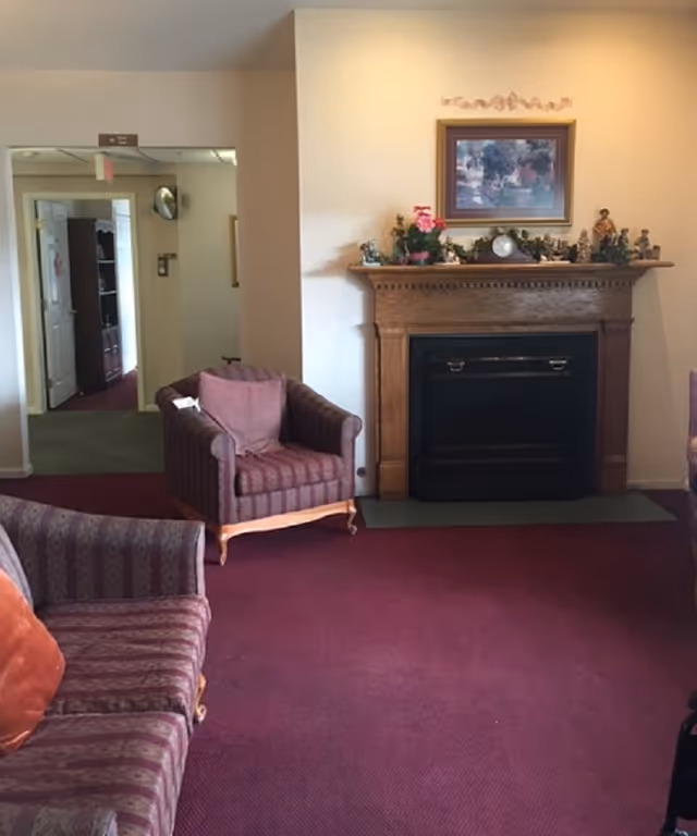A cozy living room area with a maroon carpet, two upholstered armchairs, and a matching sofa. There is a wooden fireplace mantel decorated with small figurines, a clock, and a framed painting above it. The room has light-colored walls and an open doorway leading to another area of the facility.