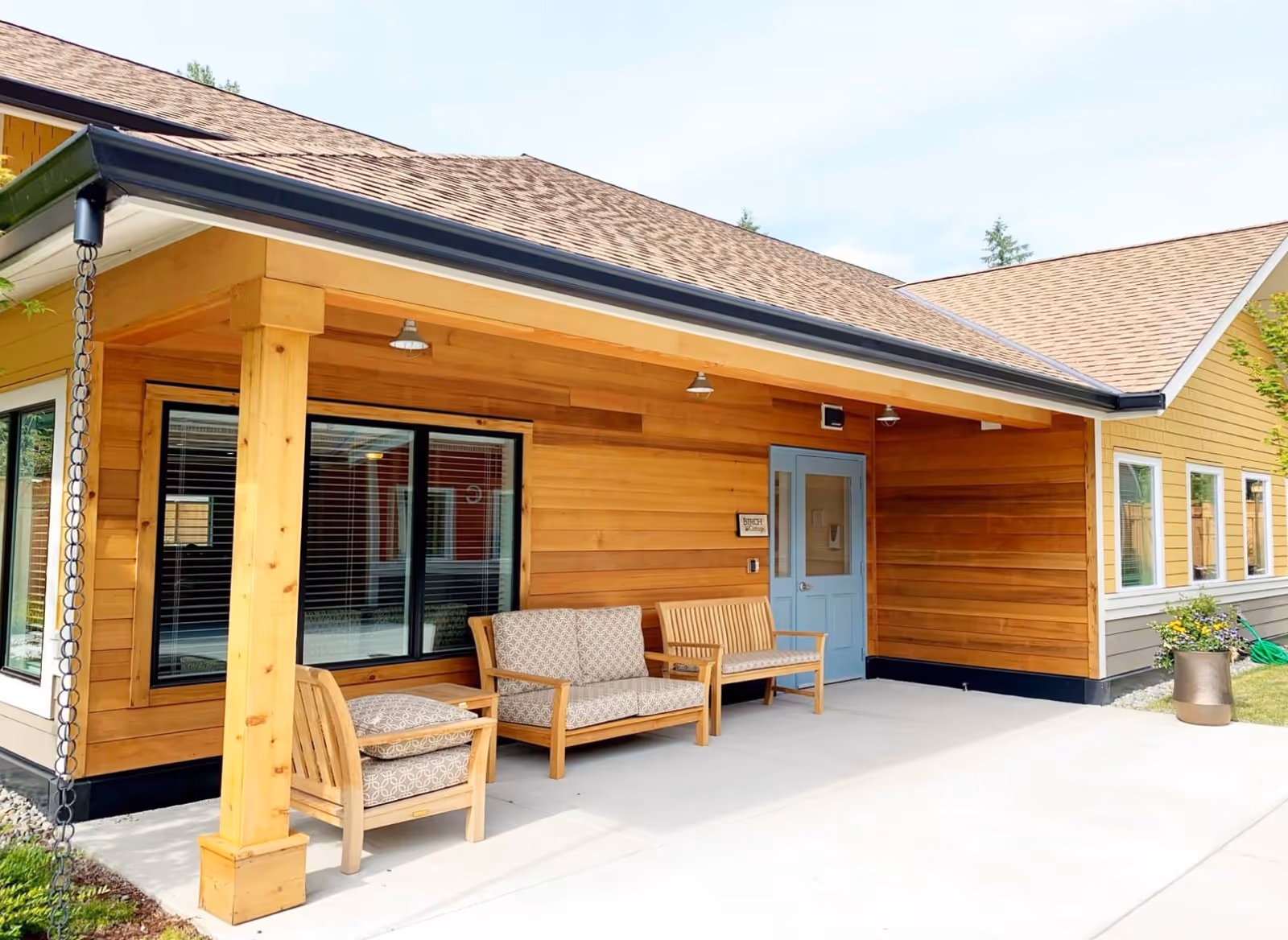 Covered outdoor seating area at Cottages of Renton with wooden chairs and a loveseat with patterned cushions, a blue door, and wood-paneled walls under a shingled roof.