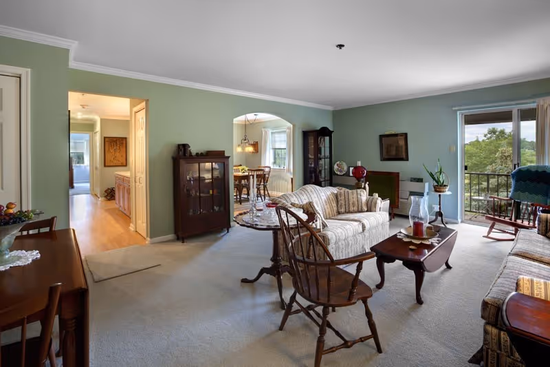 A spacious living room with light green walls and beige carpet. The room features a striped sofa, wooden chairs, a coffee table with decorative items, a glass-front cabinet, and a side table with a plant. There is a sliding glass door leading to a balcony with a view of trees outside. An archway leads to a dining area with a wooden dining table and chairs, and a hallway with wooden flooring is visible to the left.