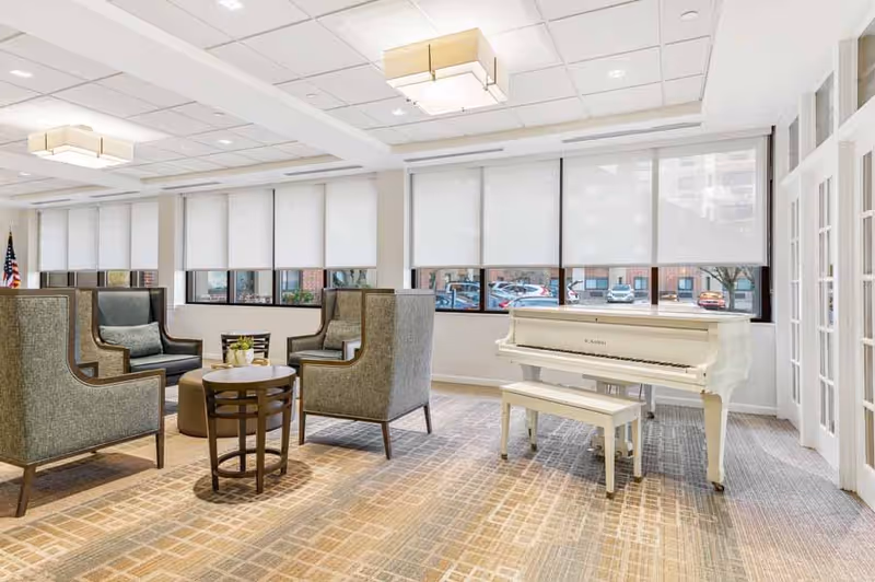 A bright and spacious common area with four high-backed armchairs arranged around a small round table. A white grand piano with a matching bench is positioned near large windows with white roller shades. The room has a patterned carpet, white walls, and ceiling lights, creating a welcoming and comfortable atmosphere.