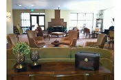 A spacious living room area with multiple striped armchairs arranged around a central fireplace. Large windows allow natural light to fill the room. A wooden table with decorative items, including a plant and a small chest, is visible in the foreground.