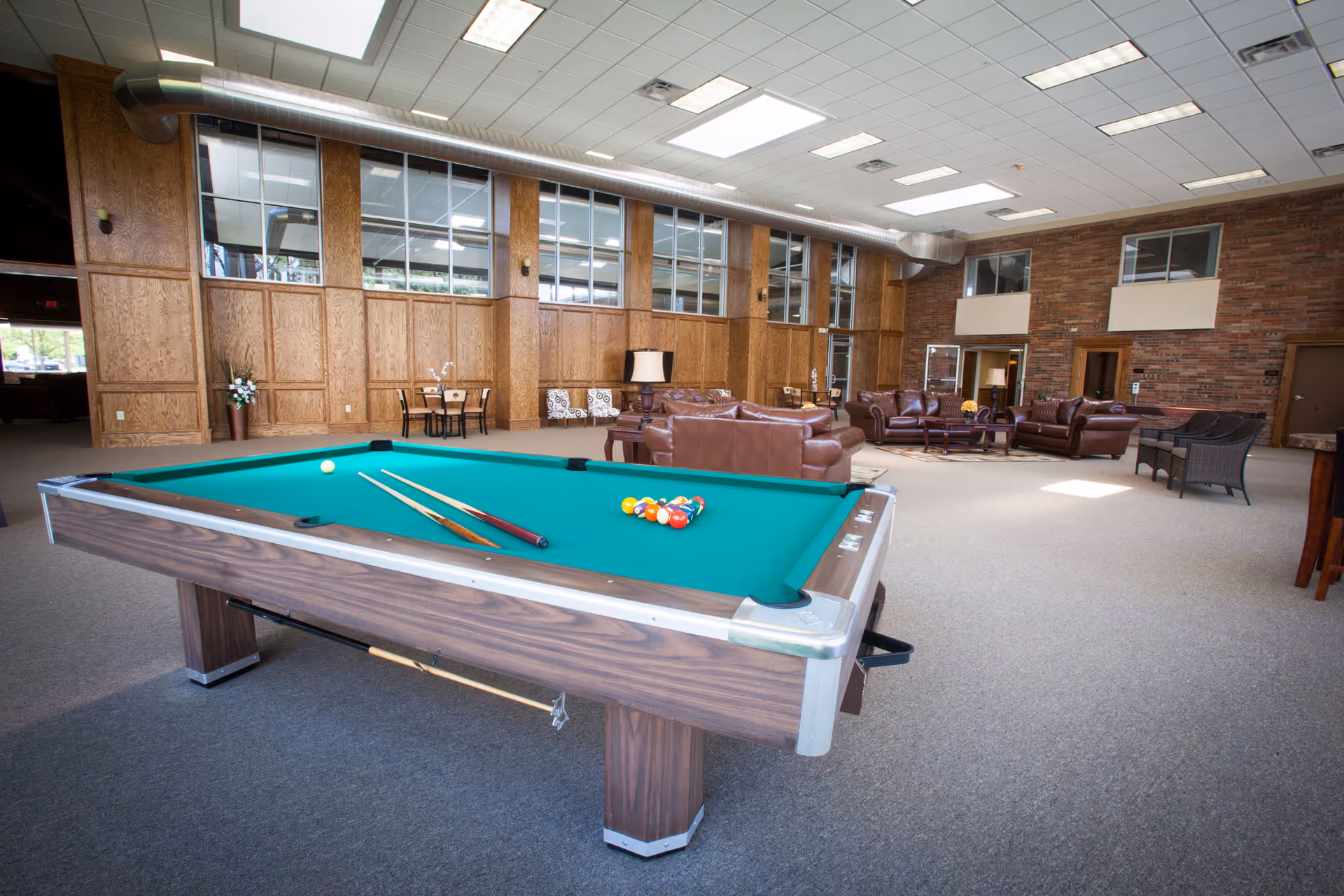 Open community lounge with a pool table in the foreground and leather sofas and seating areas under high ceilings.