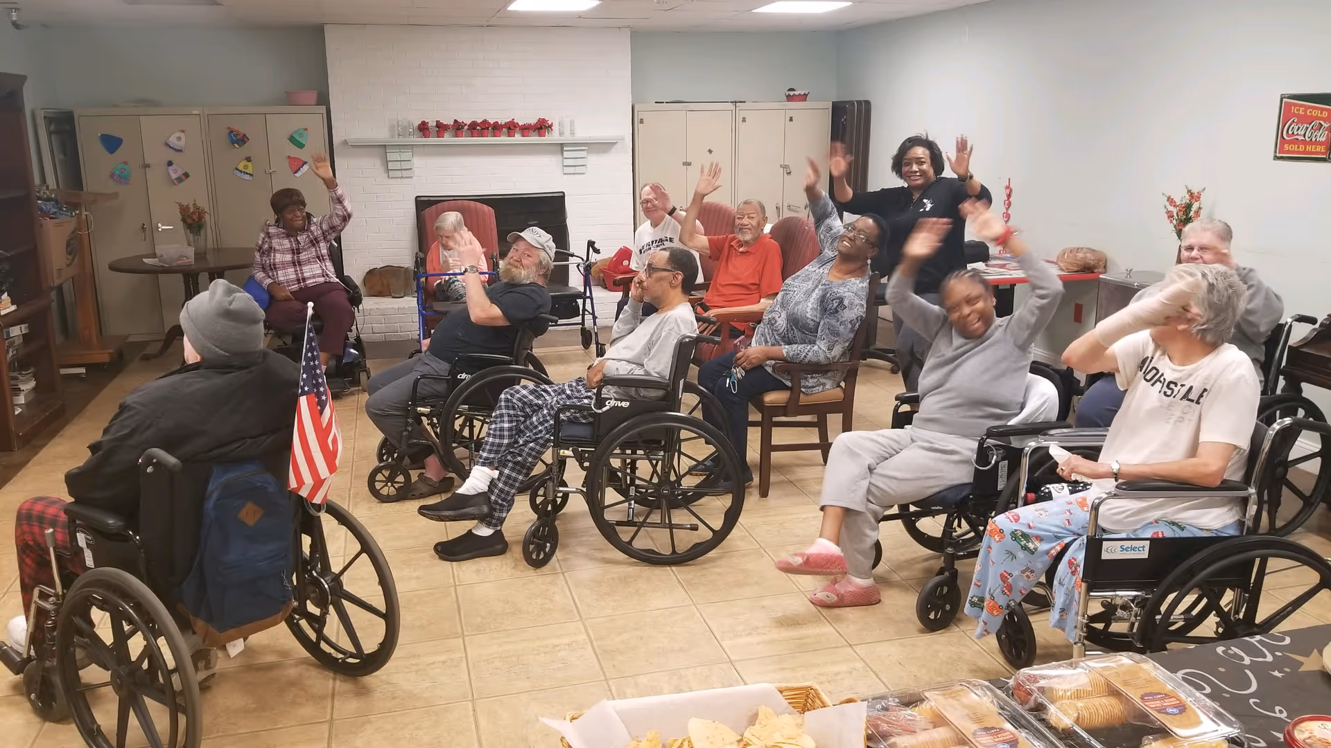 A group of elderly people, some in wheelchairs, sitting in a common room with a fireplace. They are smiling and waving at the camera. A staff member is standing among them, also waving. The room has tiled floors, cabinets, and a table with snacks in the foreground.