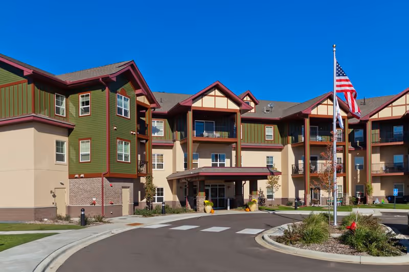 Exterior view of a multi-story senior living facility with green and beige walls, red trim, balconies, and an entrance canopy. An American flag is flying on a flagpole in front of the building under a clear blue sky.
