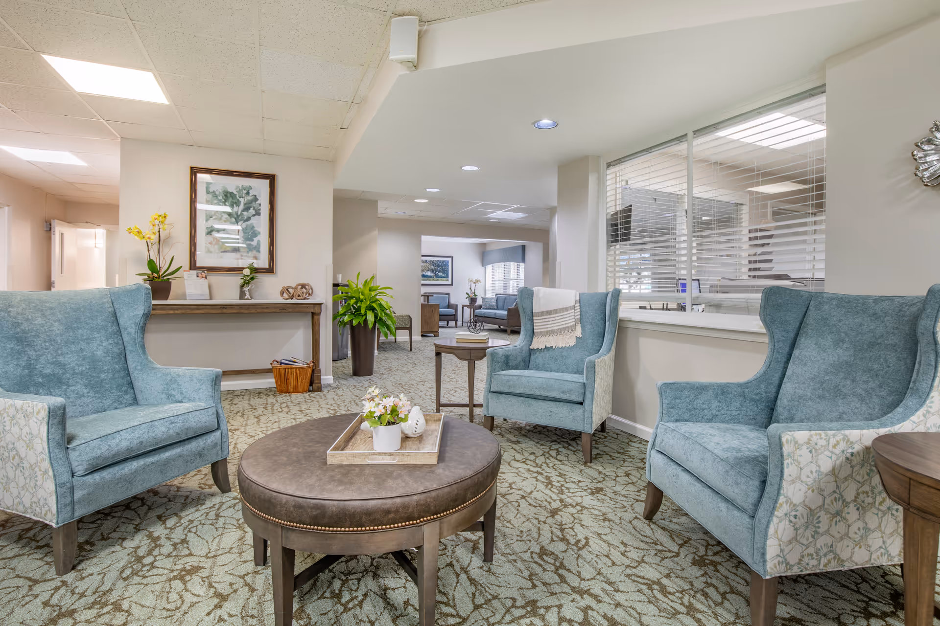 A cozy seating area in a senior living facility with three blue upholstered armchairs arranged around a round brown ottoman with a tray holding a small flower pot. The room has patterned carpet, a console table with plants and framed artwork on the wall, and a window with blinds looking into an office area.