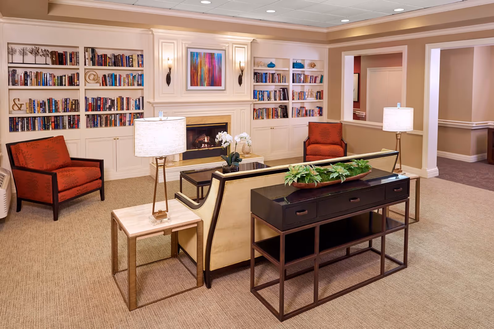A cozy living room area in an assisted living facility featuring a beige sofa with a black frame, two red armchairs, a coffee table, and two side tables with lamps. Behind the sofa is a console table with a decorative plant. The room has built-in white bookshelves filled with books on either side of a fireplace with a colorful abstract painting above it. The walls are painted in neutral tones and the floor is carpeted.