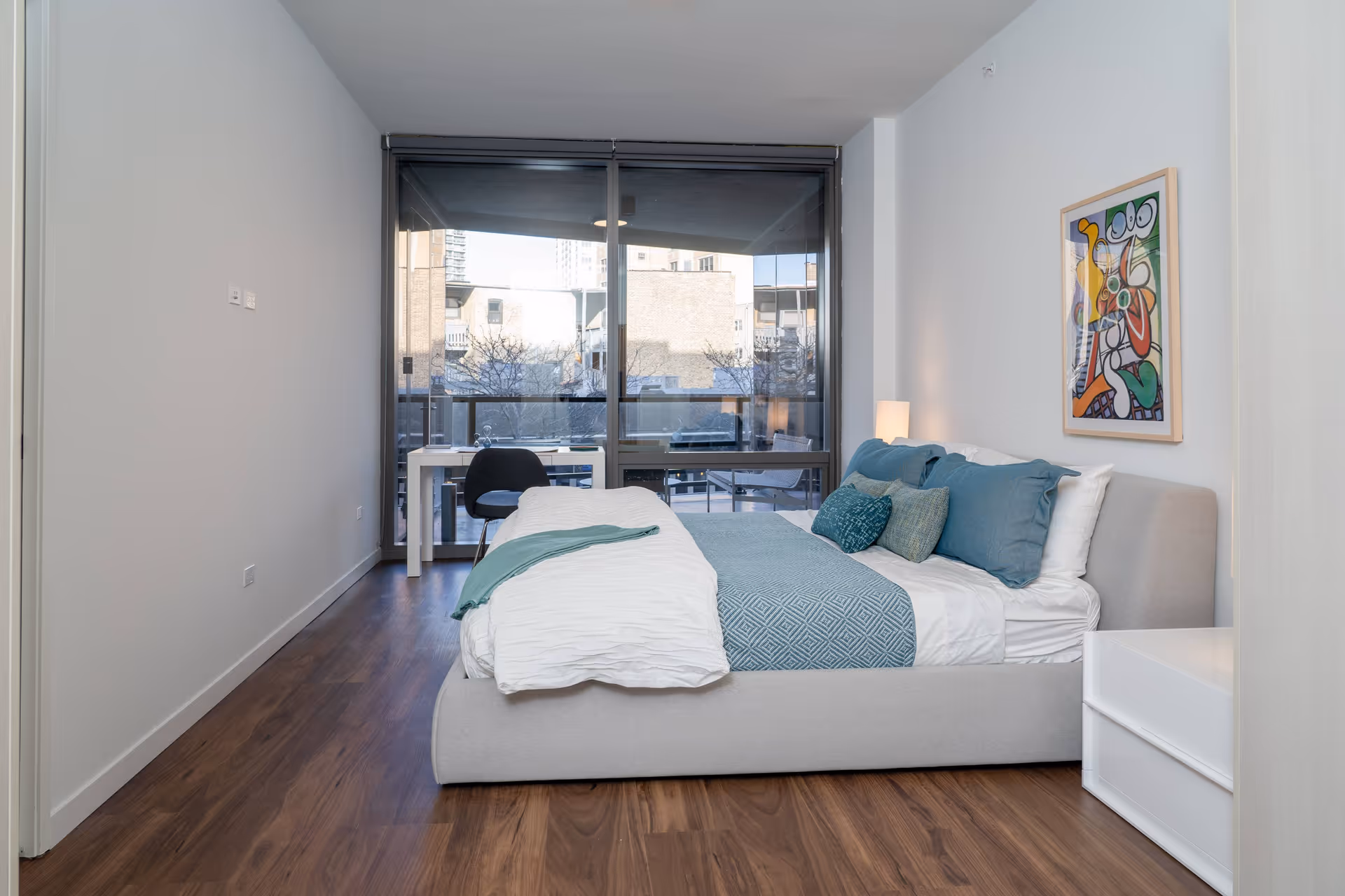 A modern bedroom with a large bed featuring white and teal bedding and multiple pillows. There is a white nightstand with a lamp on the right side of the bed. A colorful abstract painting hangs on the wall above the bed. At the far end of the room, there is a large window with a sliding glass door leading to a balcony with outdoor seating. A small white desk and a black chair are positioned near the window. The room has wooden flooring and white walls.