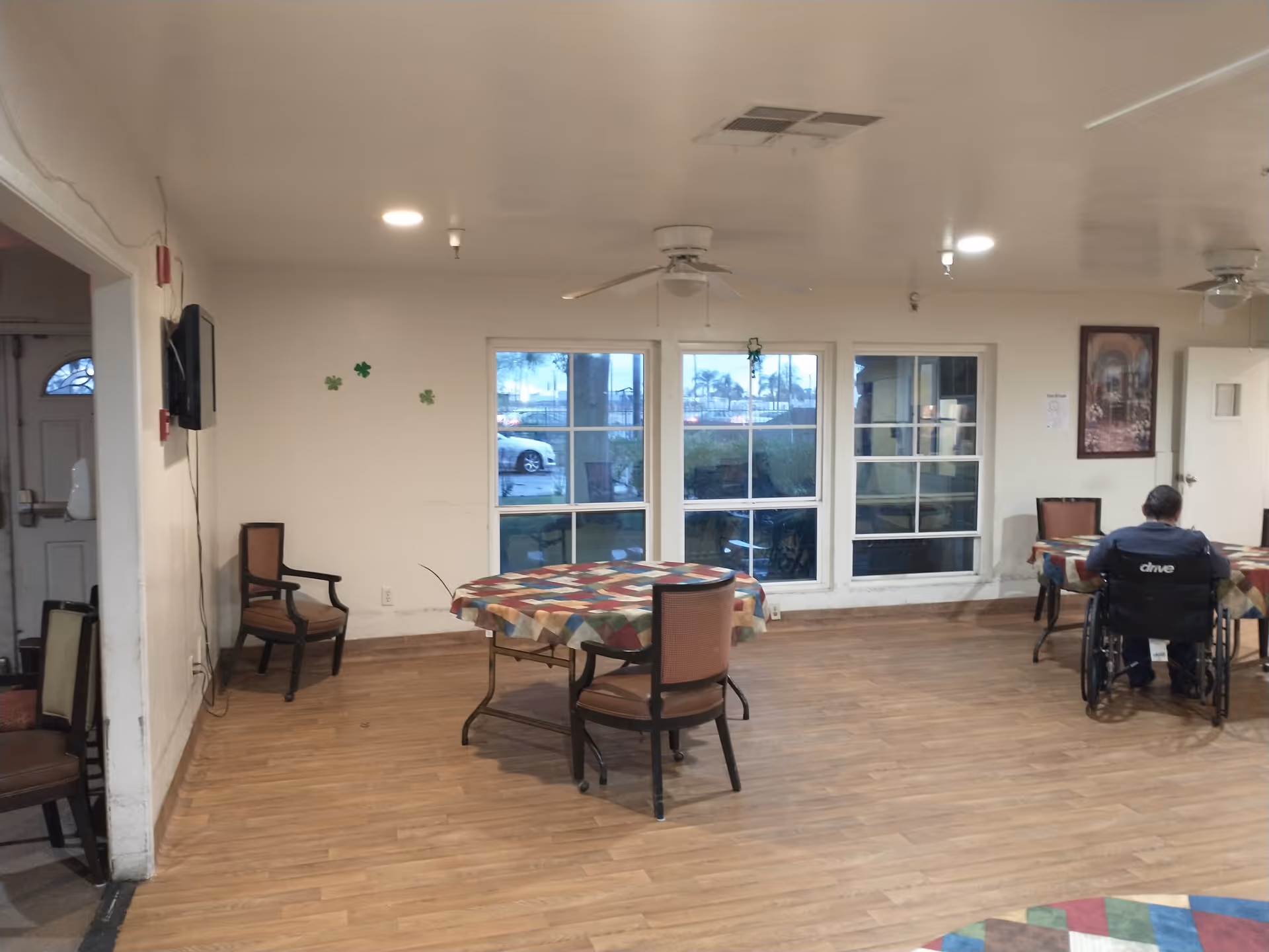 Interior view of a common area in an assisted living facility with wooden flooring, round tables covered with colorful checkered tablecloths, chairs around the tables, large windows showing an outside view, ceiling fans, and a person in a wheelchair sitting at one of the tables.