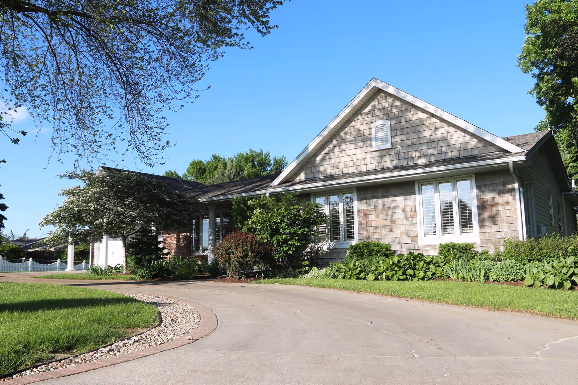 Single-story residential-style building with a curved driveway, manicured lawn and shrubs under a clear blue sky.
