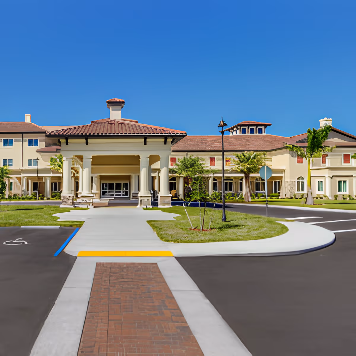 Front exterior view of a senior living facility with a covered entrance, beige walls, red-tiled roof, and a clear blue sky. There is a paved walkway leading to the entrance, surrounded by green grass and small trees.