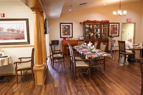 Interior view of a dining room in a senior living facility with wooden floors, several tables and chairs, a china cabinet decorated with fall-themed garlands, framed artwork on the walls, and a senior woman sitting at a table on the right side.