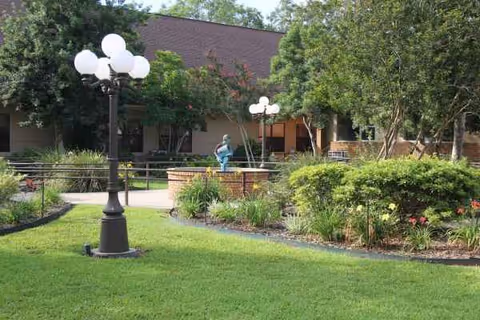 Landscaped outdoor courtyard with grassy lawn, lamp posts, shrubs and a small fountain or statue in front of the facility building.