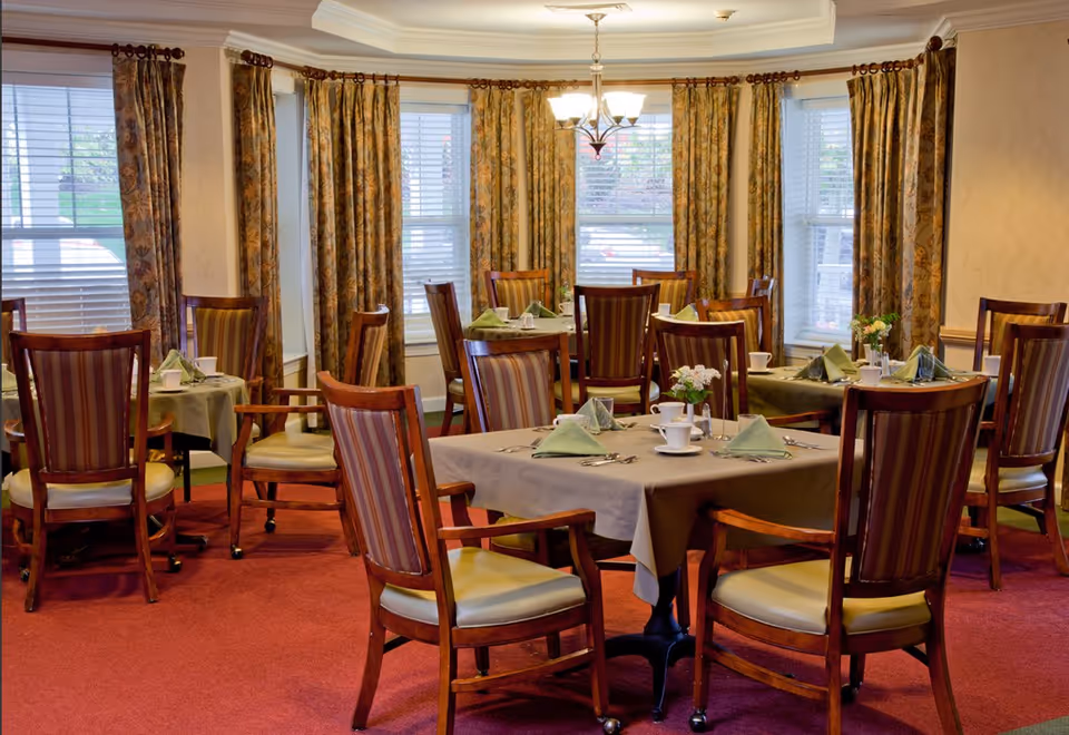 Dining room with several tables set with napkins, teacups and wooden chairs in front of bay windows and patterned curtains.