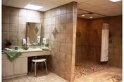 A tiled bathroom area in a nursing and rehabilitation center featuring a large mirror above a sink with two faucets, a small stool, and a walk-in shower with a white curtain. The walls and floor are covered in beige and brown tiles.