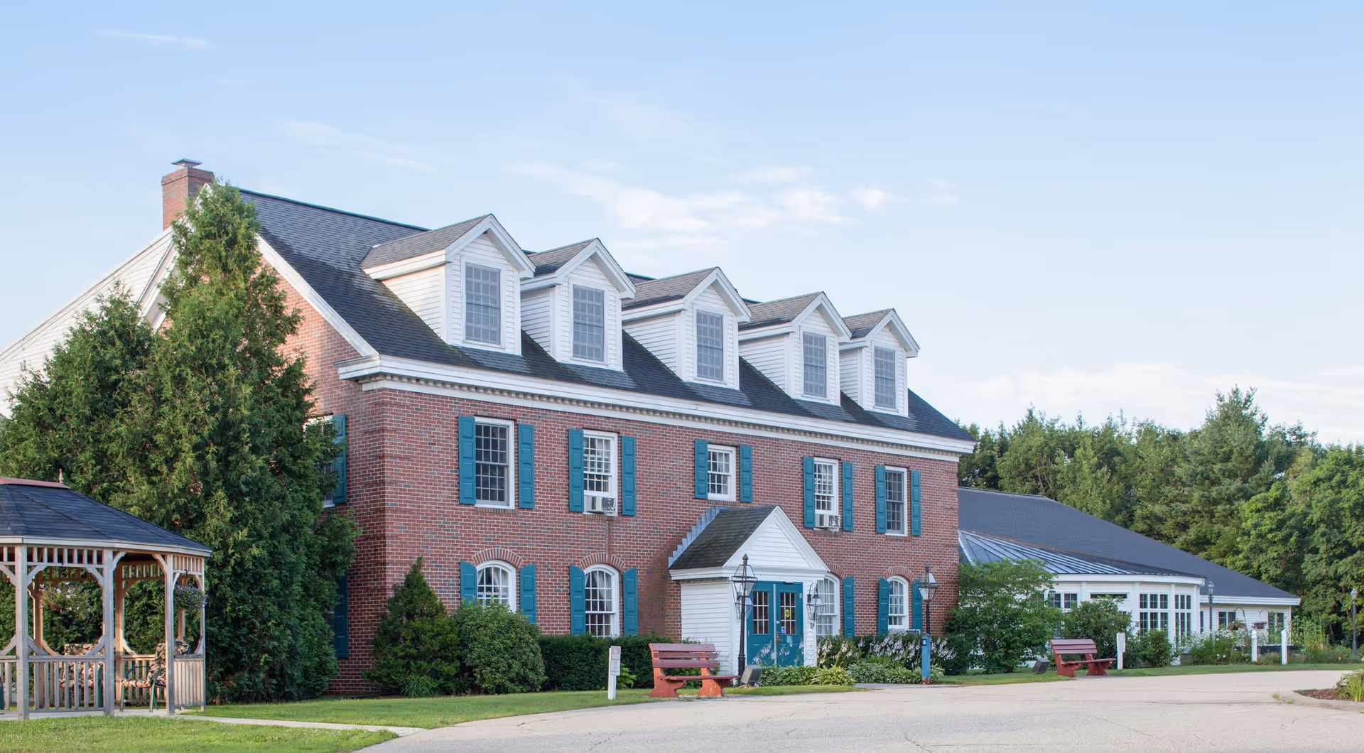 Red-brick colonial-style building with dormer windows and green shutters, a gazebo to the left, benches and landscaped grounds in front.