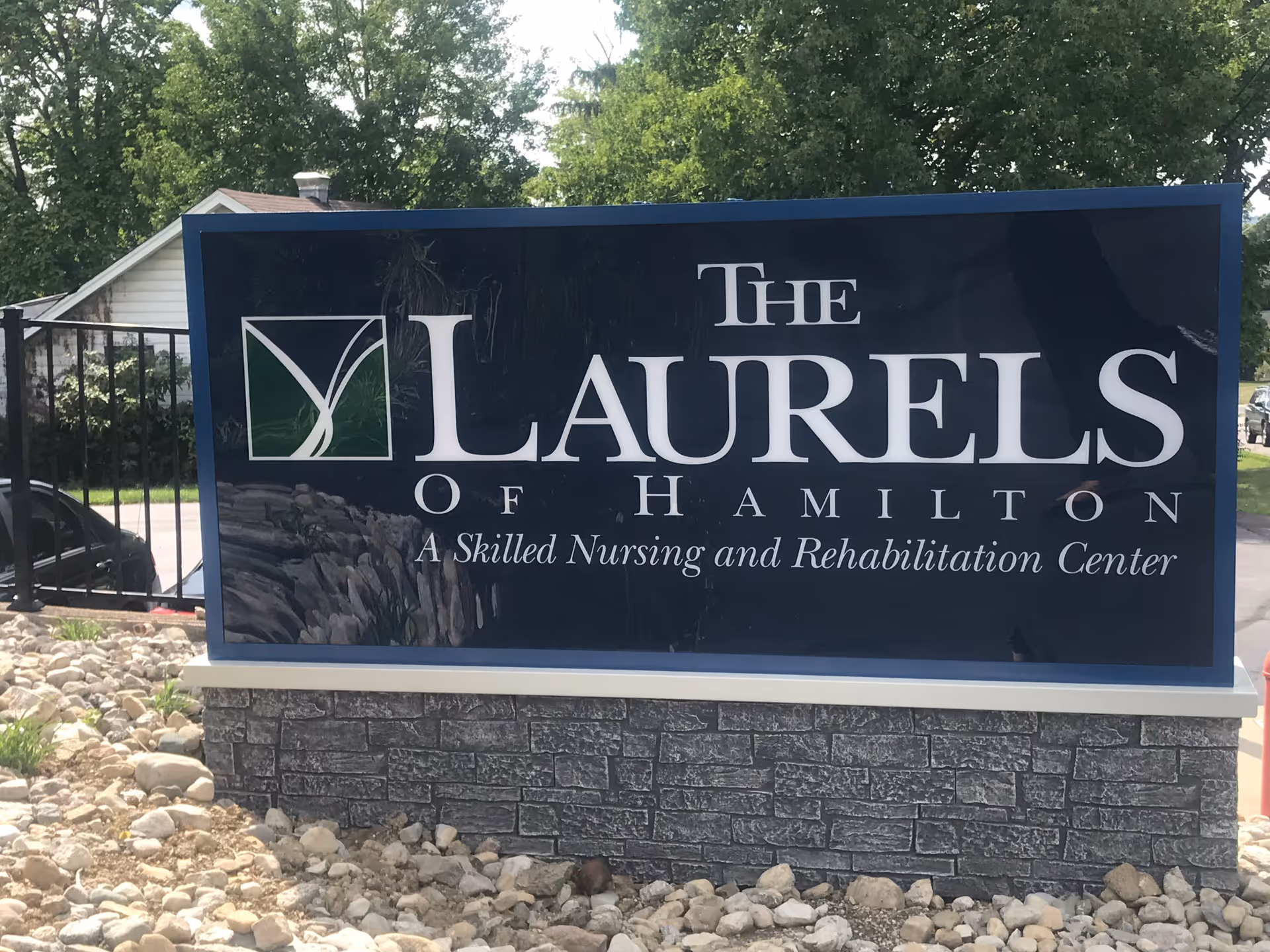 Outdoor sign for The Laurels of Hamilton, a skilled nursing and rehabilitation center, surrounded by rocks and greenery with trees and a house in the background.