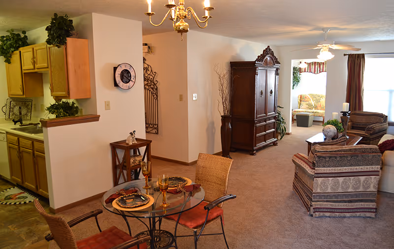 Interior view of a senior living facility showing a combined living and dining area. The dining area has a round glass table set with plates and glasses, surrounded by four chairs with red cushions. To the left is a kitchen with wooden cabinets and a sink. The living area features patterned upholstered chairs, a wooden armoire, a coffee table with decorative items, and a ceiling fan. There is a window with curtains and a small seating area visible in the background.