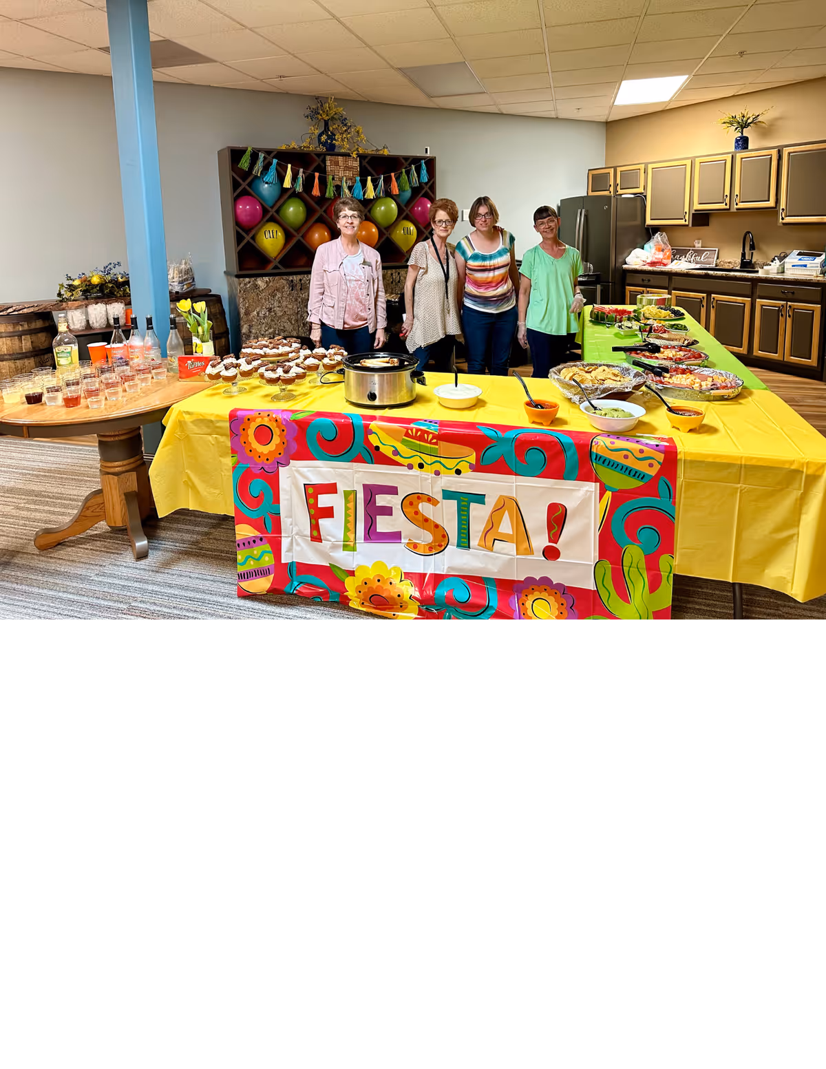 Four women standing behind a table covered with a yellow tablecloth and a colorful banner that reads 'FIESTA!'. The table is set with various food dishes, cupcakes, and a slow cooker. Behind them is a kitchen area with cabinets and a refrigerator, and a decorative shelf with colorful balloons and tassels.