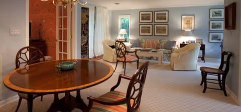 Open living and dining area with a round wooden dining table and chairs in the foreground and a cozy seating area with sofas, lamps, and framed artwork in the background.