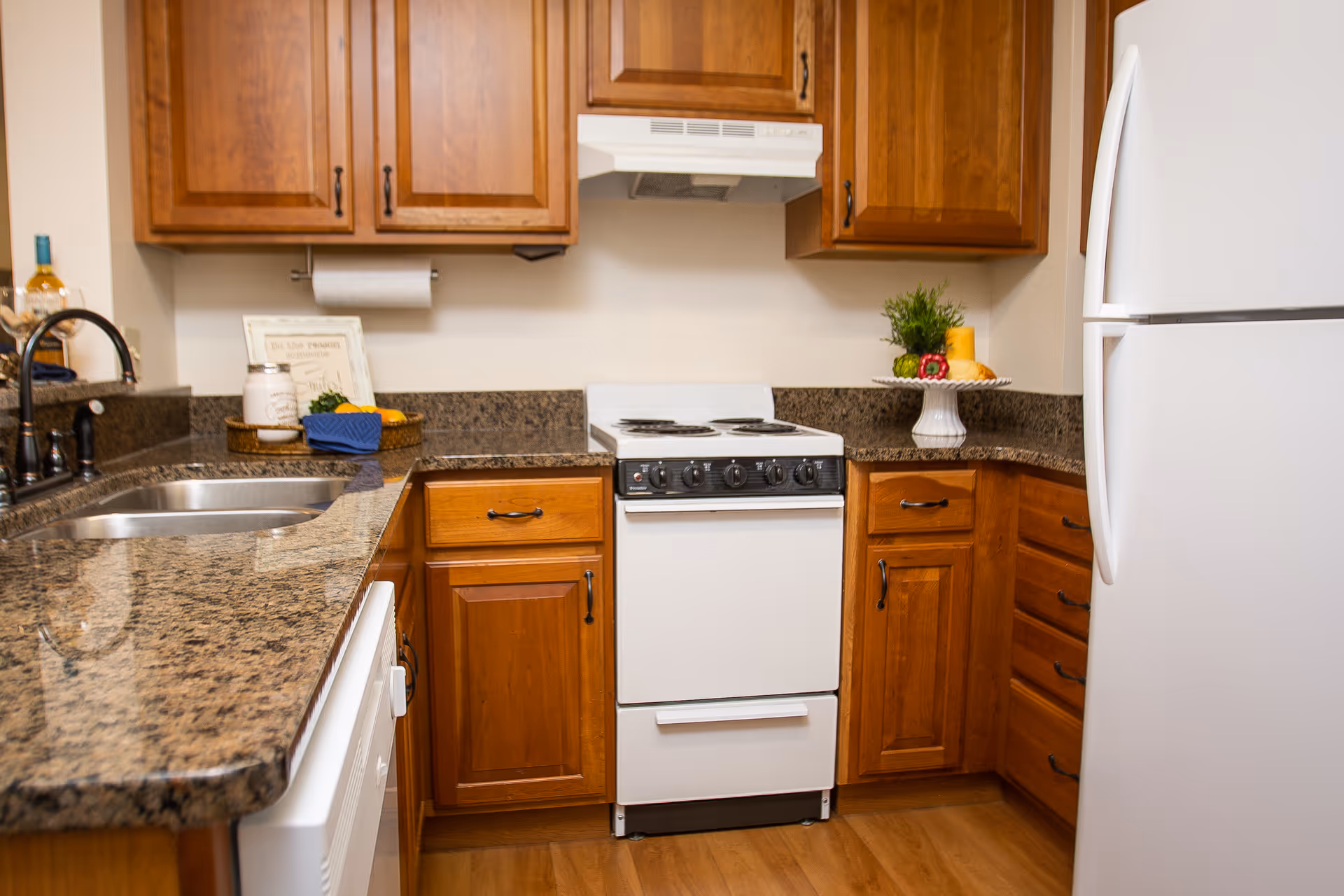 A compact kitchen with wooden cabinets, granite countertops, a white electric stove with four burners, a white refrigerator, and a stainless steel sink. The countertop has decorative items including a small plant, a fruit basket, and a framed sign.