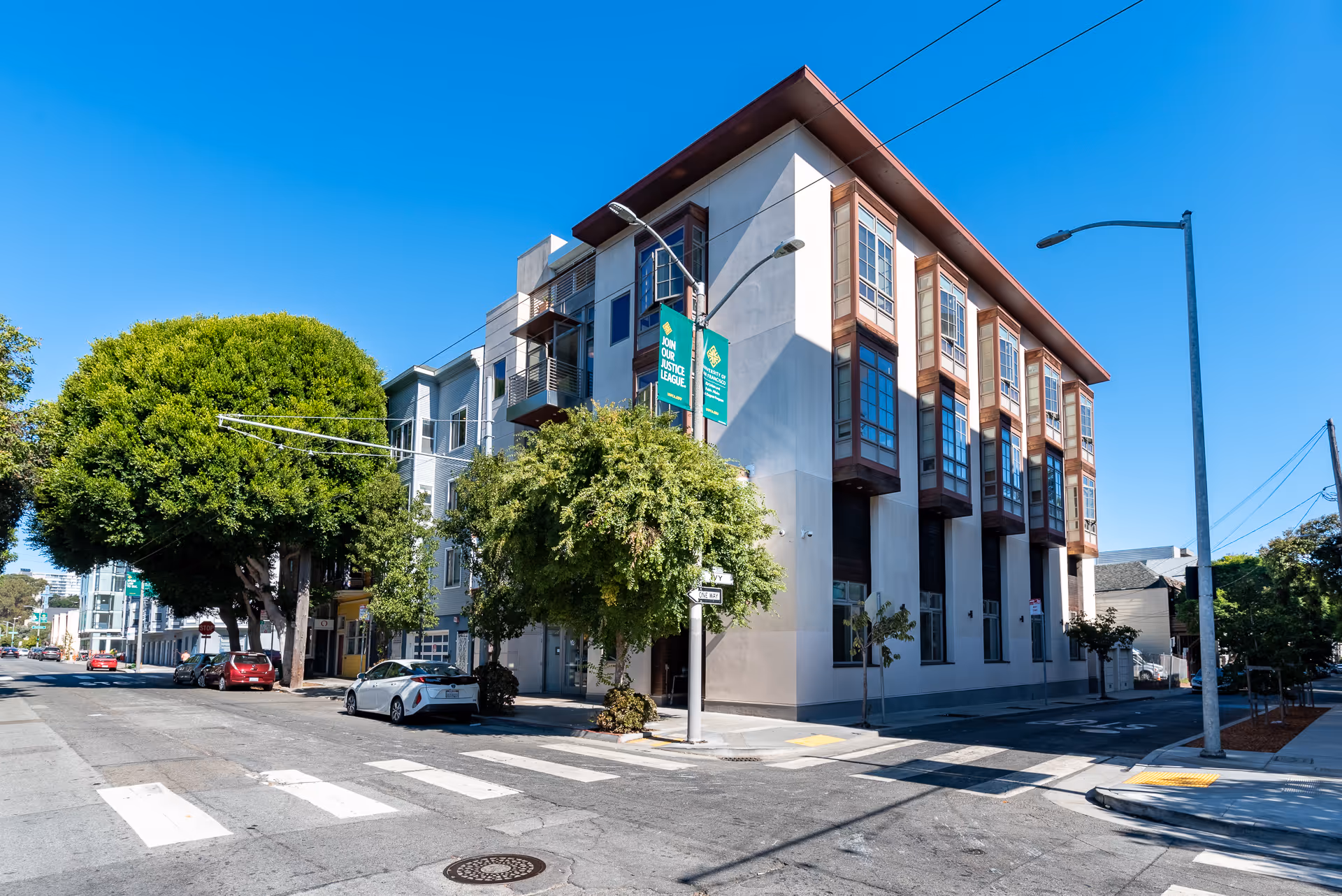 Street view of a modern multi-story building on a sunny day with clear blue sky. The building has large windows and is located at a street corner with trees and parked cars nearby.