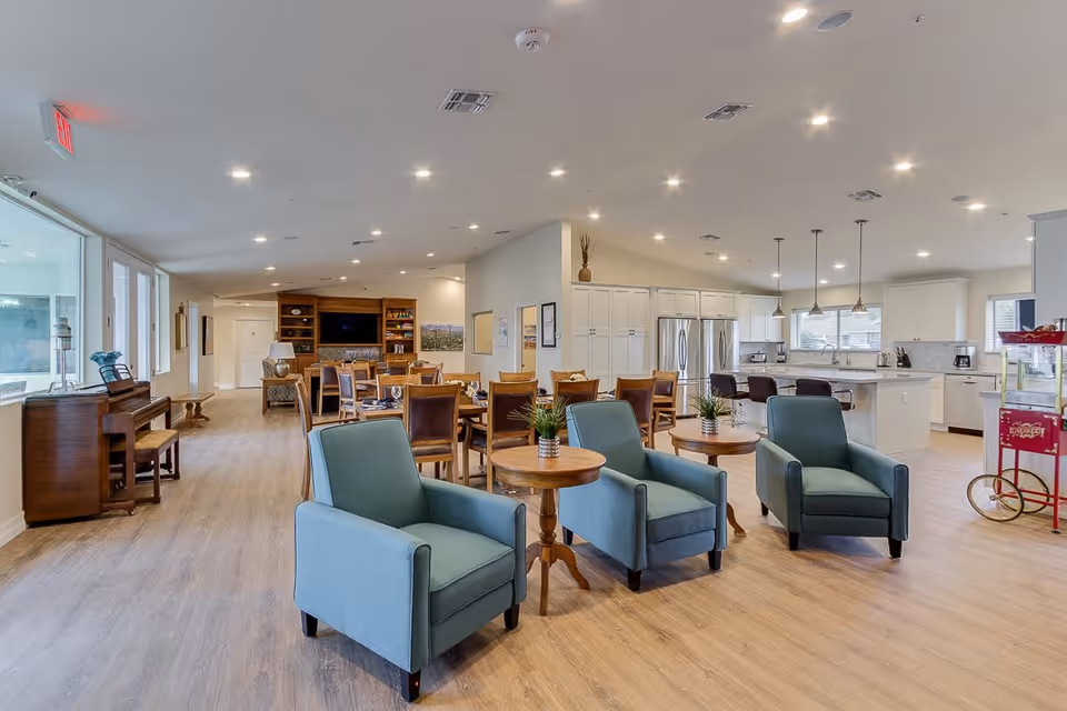 A spacious common area featuring comfortable blue chairs, wooden tables, and a dining setup with a kitchen in the background. Large windows allow natural light to fill the room.