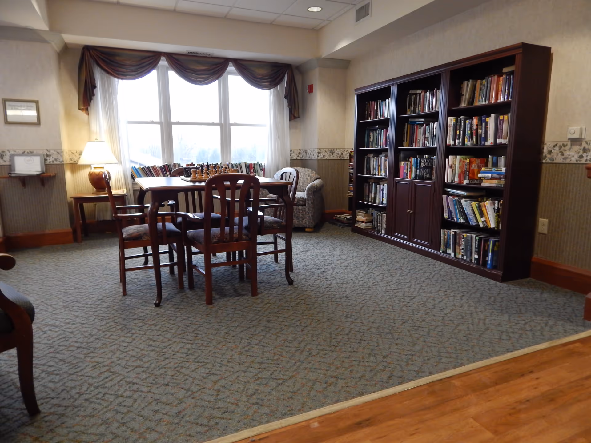 A cozy reading and game area in a senior living facility with a wooden table and four chairs, a chess set on the table, a large bookshelf filled with books, a comfortable armchair, a lamp on a side table, and large windows with draped curtains letting in natural light.