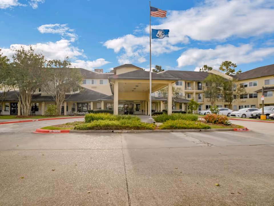 Front exterior view of Whealdon Estates by Barclay House, showing a large covered entrance with two flagpoles flying the American flag and a state flag. The building is a multi-story structure with beige walls and dark roofing, surrounded by trees, shrubs, and parked cars under a partly cloudy sky.
