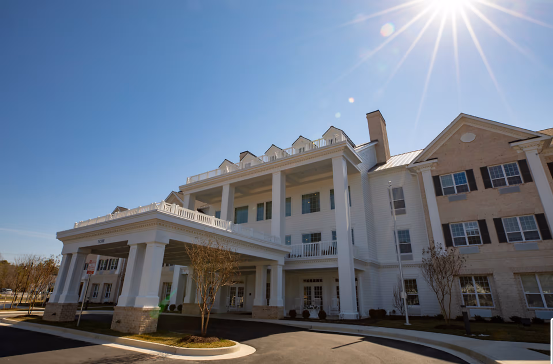 Front exterior of a senior living facility with a covered portico, tall white columns, and multiple windows under a bright sun.
