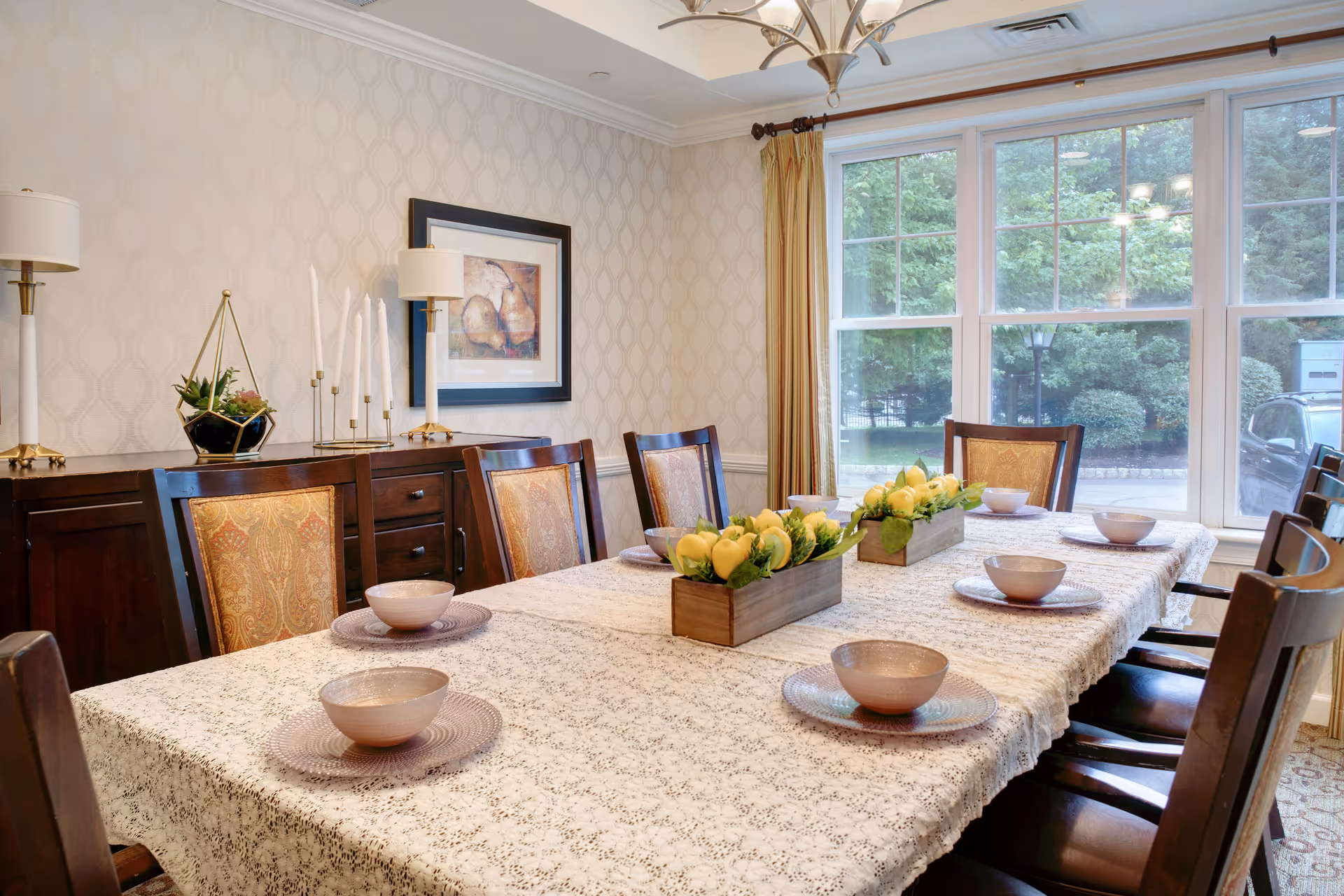 A dining room with a long table covered by a white patterned tablecloth, set with bowls and plates. The table has two rectangular wooden planters filled with yellow tulips as centerpieces. The room features wooden chairs with upholstered backs, a sideboard with lamps, candles, and a framed artwork on the wall. Large windows with beige curtains show greenery and a parked car outside.