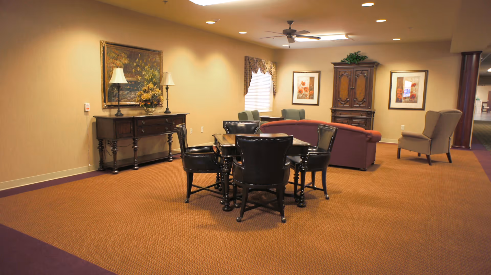 A cozy common area with a square wooden table surrounded by four black leather chairs in the foreground. Behind the table, there is a red sofa, two green armchairs near a window with blinds and valance, and a beige wingback chair. The walls are decorated with framed floral artwork and a large painting above a dark wooden sideboard with two lamps and a floral arrangement. The room has warm lighting and a ceiling fan.
