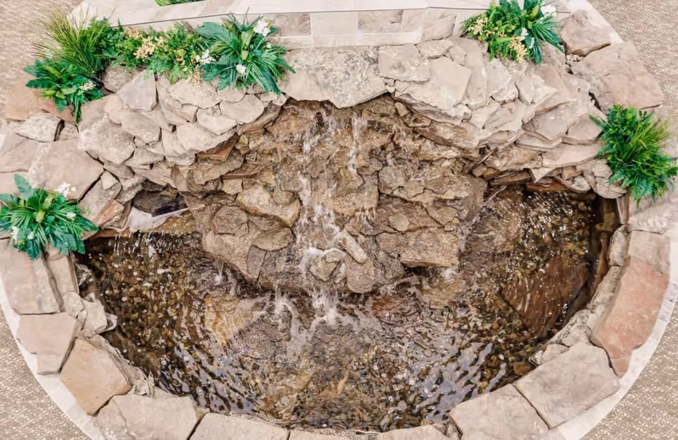 Top-down view of a small indoor water feature with flowing water over rocks, surrounded by stone edging and green plants with white flowers.