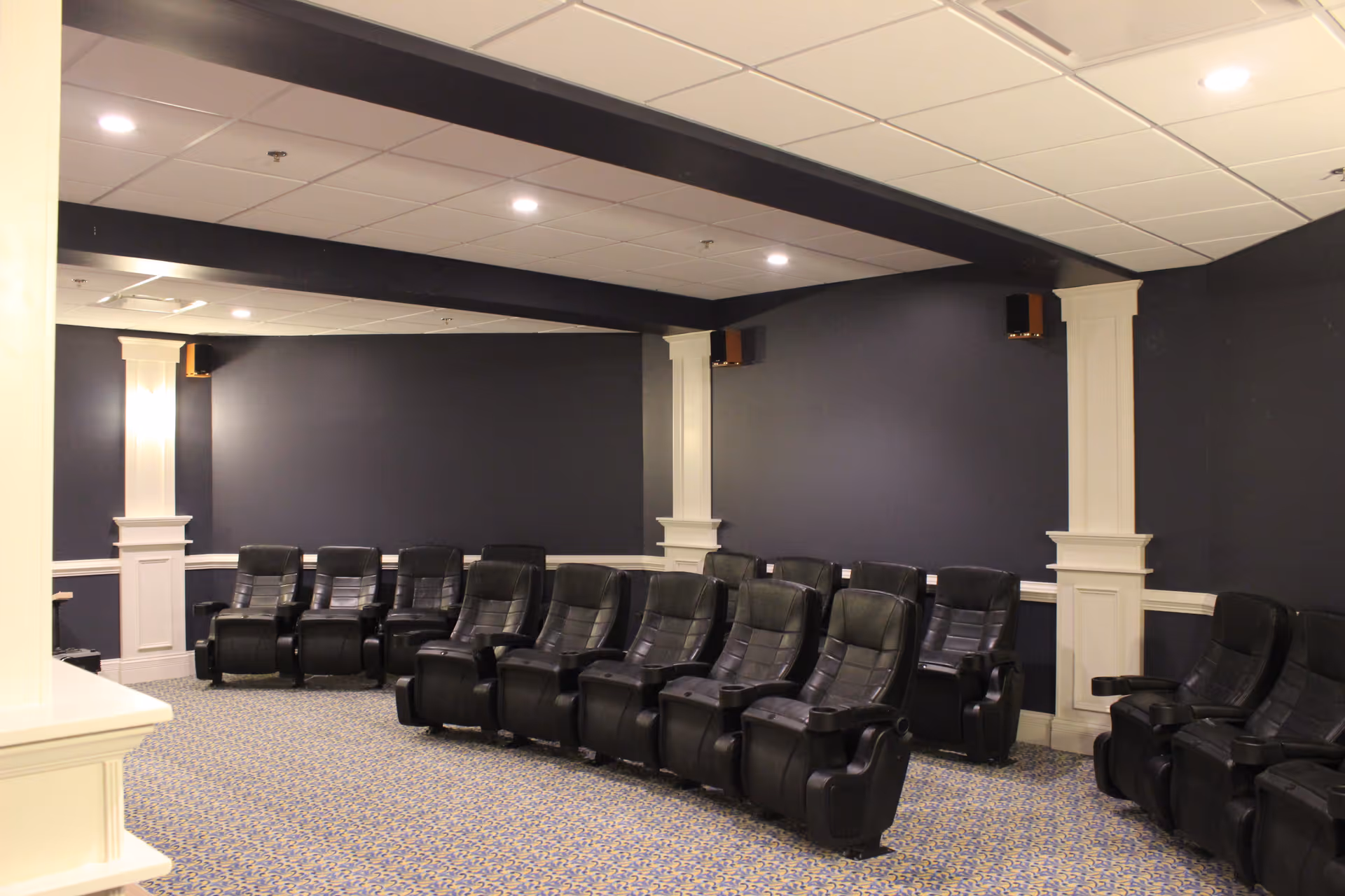 A room with multiple black leather recliner chairs arranged in rows facing the same direction, with dark blue walls, white pillars, and a patterned carpeted floor, likely a small theater or media room.