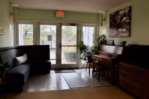 A cozy interior common area with a black cushioned bench along the left wall, a piano with a bench on the right, a wooden dresser with a TV on top, and a large windowed door leading outside. There is a plant next to the piano and a painting of trees hanging above it.