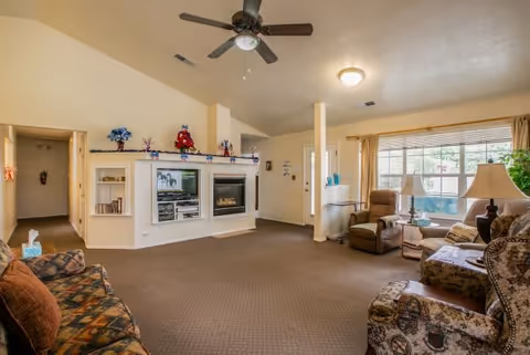 A spacious living room with a ceiling fan, carpeted floor, and a large window letting in natural light. The room features a built-in entertainment center with a TV and fireplace, decorated with holiday ornaments. There are multiple upholstered chairs and sofas arranged around the room, along with side tables and lamps.