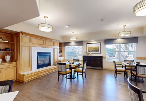 A cozy common area with wooden flooring, several round tables with chairs, a fireplace with a wooden mantel, built-in shelves with decorative items, large windows letting in natural light, and a sideboard with a framed picture above it.