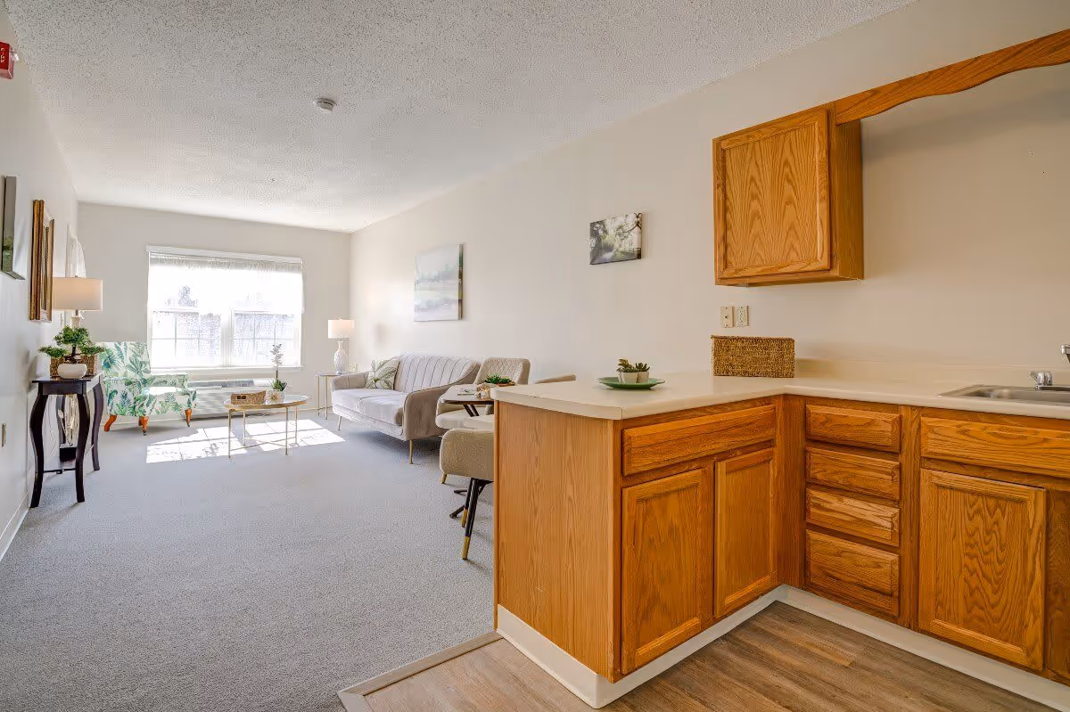 A bright living area with a large window letting in natural light. The room features a beige sofa, a patterned armchair, a coffee table, and side tables with lamps. Adjacent to the living area is a small kitchen with wooden cabinets, a countertop, and a sink. The walls are light-colored and decorated with a few framed pictures.
