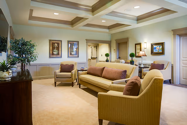A cozy living room area in an assisted living facility featuring beige carpet and walls with crown molding. The room has a yellow sofa and matching armchairs with brown cushions, a wooden cabinet with plants on top, framed floral artwork on the walls, and a table lamp on a side table. The ceiling has recessed lighting and decorative beams.