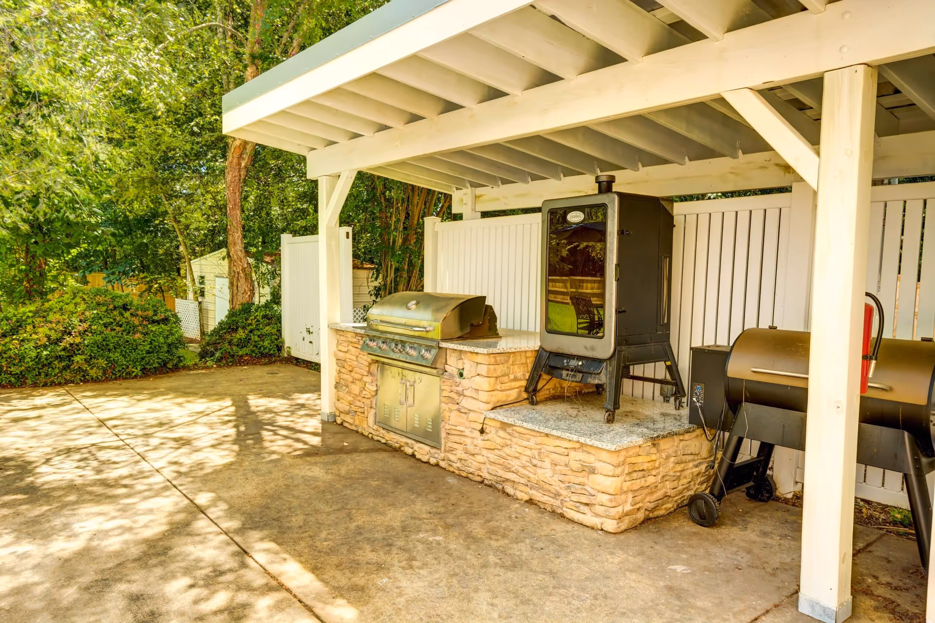 Outdoor covered grilling area with a stainless steel gas grill, a vertical smoker, and a charcoal grill, surrounded by a white wooden fence and greenery.