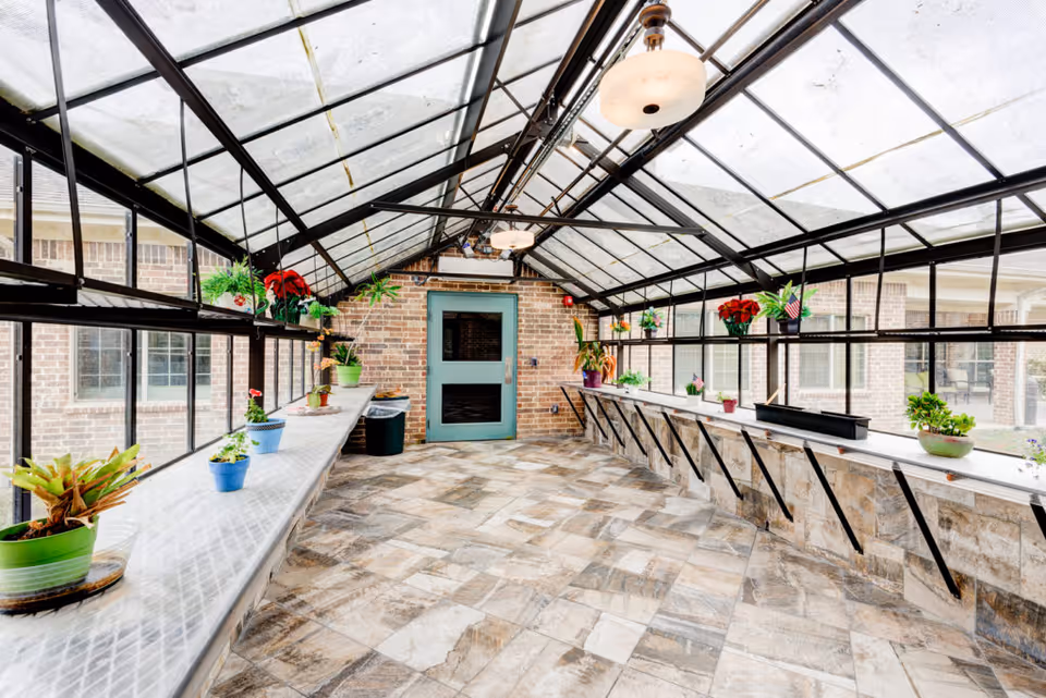 A bright sunroom with a glass ceiling and large windows on both sides. The room has a tiled floor and long narrow shelves along the windows holding various potted plants. A green door is visible at the far end of the room, and two ceiling lights hang from the glass roof structure.