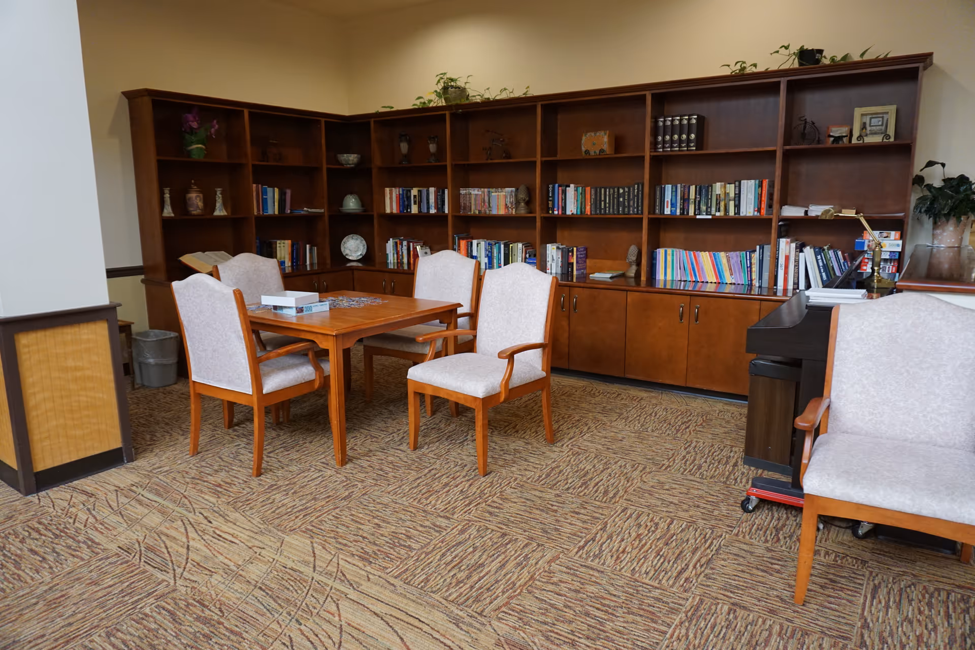 A cozy reading or activity room with a wooden table surrounded by four cushioned chairs. Behind the table is a large wooden bookshelf filled with books, decorative items, and plants. The room has carpeted flooring with a patterned design and neutral-colored walls.