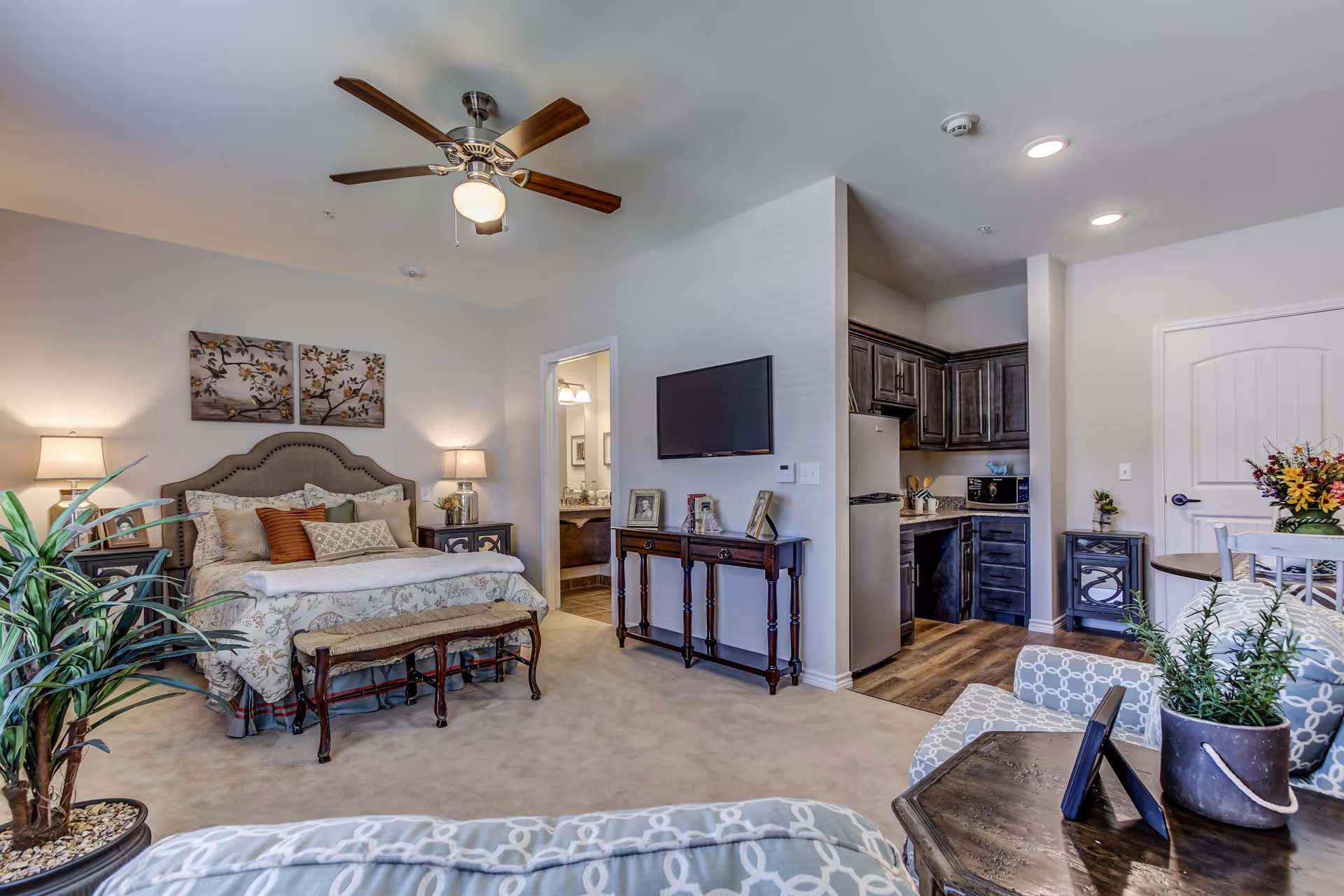 A well-lit assisted living apartment featuring a cozy bedroom with a bed, nightstands with lamps, and decorative wall art. Adjacent to the bedroom is a small kitchen area with dark cabinets, a refrigerator, and a microwave. A flat-screen TV is mounted on the wall above a wooden console table. The room also includes a sitting area with patterned armchairs, a coffee table with a potted plant, and a dining table with flowers. The bathroom is visible through an open door next to the bedroom.