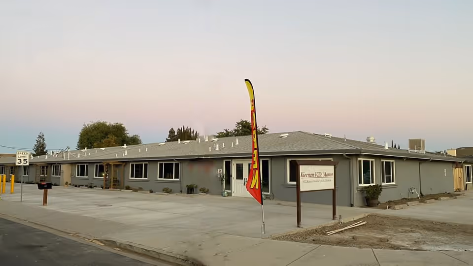 Exterior view of a single-story senior living facility building with a gray roof and walls, multiple windows, and a sign that reads 'Kiernan Ville Manor'. There is a red and yellow flag with the word 'OPEN' near the entrance, and a speed limit sign showing 35 mph on the street in front.