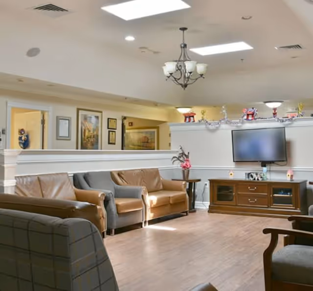 Communal lounge with leather sofas and armchairs facing a wall-mounted TV and wooden media cabinet under ceiling lights.