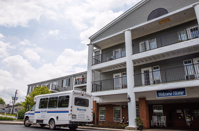 Front entrance of a senior living building with a parked Rosewalk shuttle van and a 'Welcome Home' sign.