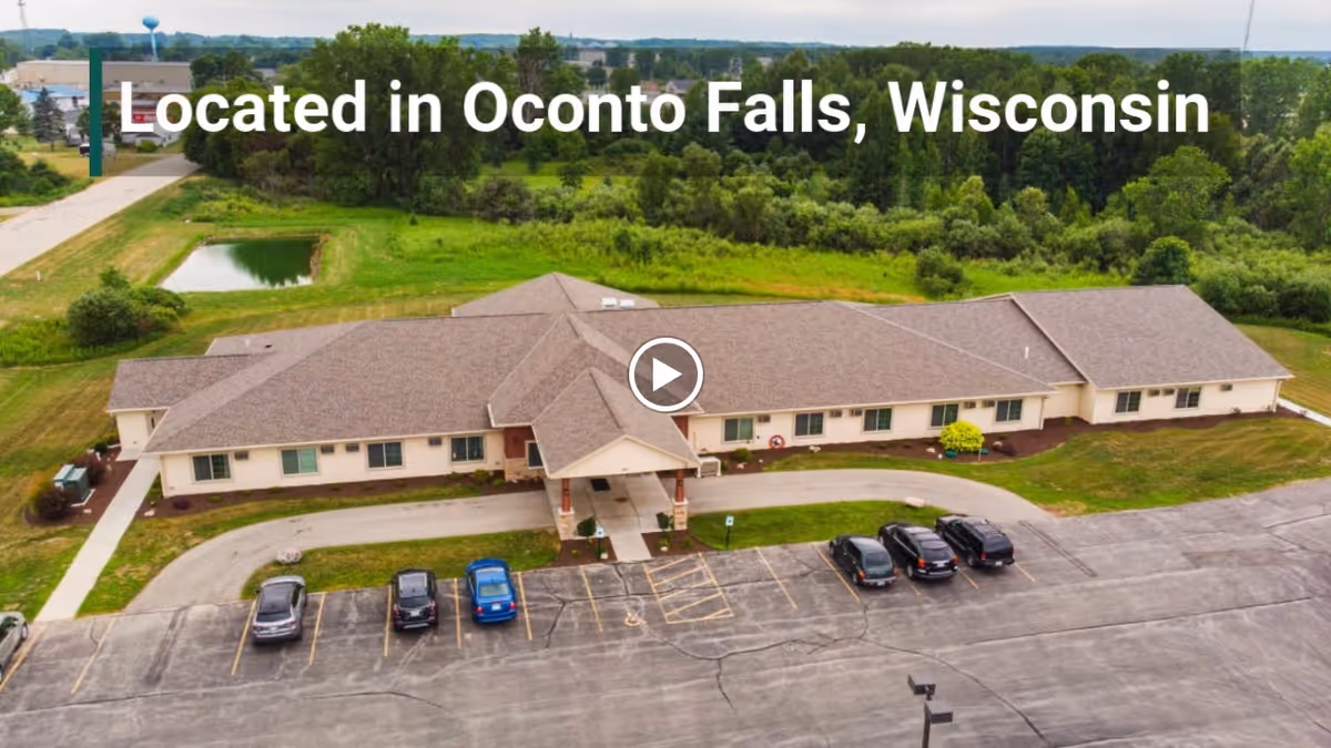 Aerial front view of a single-story memory-care building with cars in the parking lot and the overlaid text 'Located in Oconto Falls, Wisconsin'.