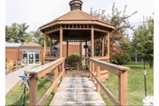 A wooden gazebo with a peaked roof at the end of a wooden walkway with railings, surrounded by green grass and trees, with a brick building visible in the background under a partly cloudy sky.