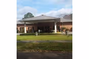 Front exterior view of a single-story brick building with a covered entrance supported by white columns. There is a well-maintained lawn and some shrubs in front of the building under a partly cloudy sky.
